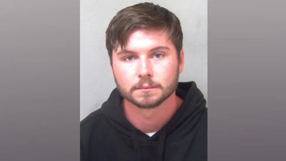 A police custody photo of Brandon Tyler. He has dark hair with a slightly floppy fringe and a short-cropped beard. He is wearing a black hoodie and looking into the camera.