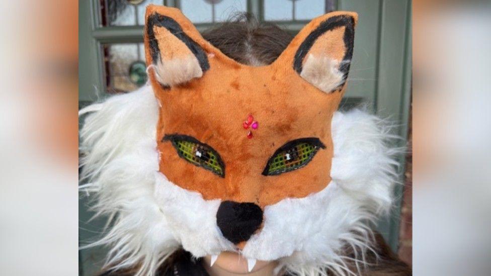 Lottie, who has long brown/blonde hair, stood in front of her front door with a fox mask, with has netting for the eyes and bushy white material around the edge of its face.