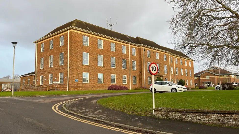 Wiltshire Police headquarters in Devizes. It is a large brown-brick building with three stories and a grass area in front. There are two white cars parked in the car park.
