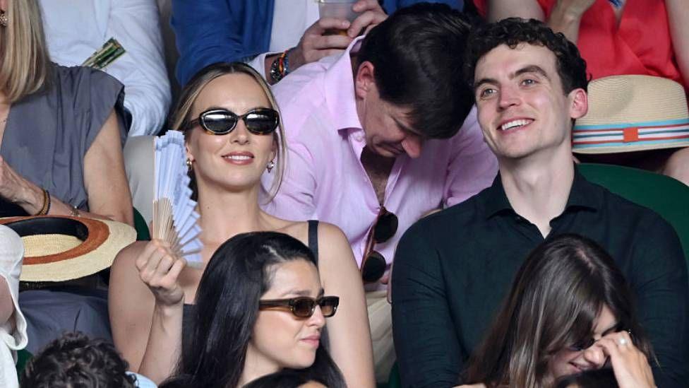 Jodie Comer sits by a man wearing a black polo shirt. Both are smiling, as Comer fans herself with a blue fan.