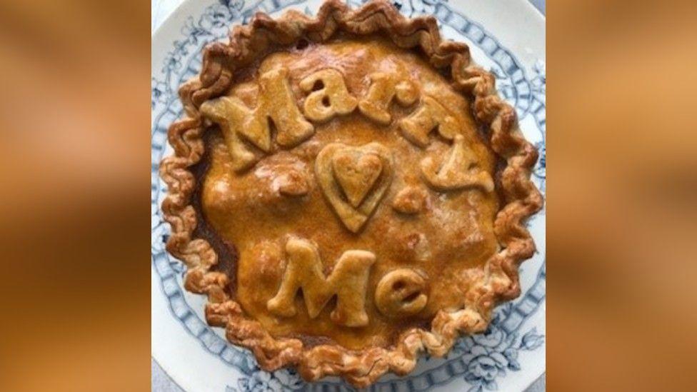 A pork pie. The pastry on top spells out "Marry Me", and there is a heart there, too. The pie is on a white and blue plate.