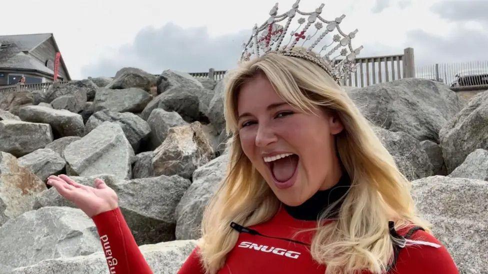 Milla Magree, with blonde shoulder length hair, wearing a read wet suit and her Miss England pageant crown. She is posing with her right hand toward the pile of rocks that are behind her. 