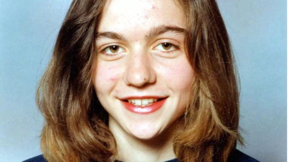 Photo shows a young Michelle Bettles, possibly while at school. She has brown eyes and shoulder-length brown hair and is smiling at the camera. There is a blue background behind her.