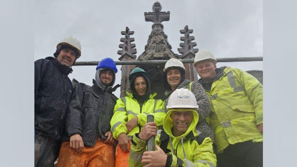 A team of five stonemasons in high visibility protective clothing and helmets smiling at the camera. Parts of Lowther Castle's ornamented pinnacle are visible in the background against a grey sky. 