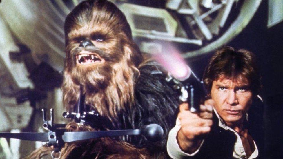 Chewbacca, a tall alien covered in brown fur, holding his bowcaster, which is a black gun that looks like a crossbow. He is standing next to Harrison Ford as Han Solo, who is firing a pistol.