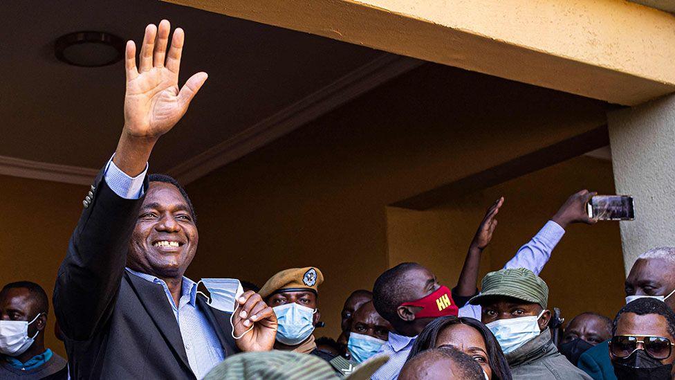 Hakainde Hichilema in an open blue shirt and dark blazer smiles and waves at his supporters.