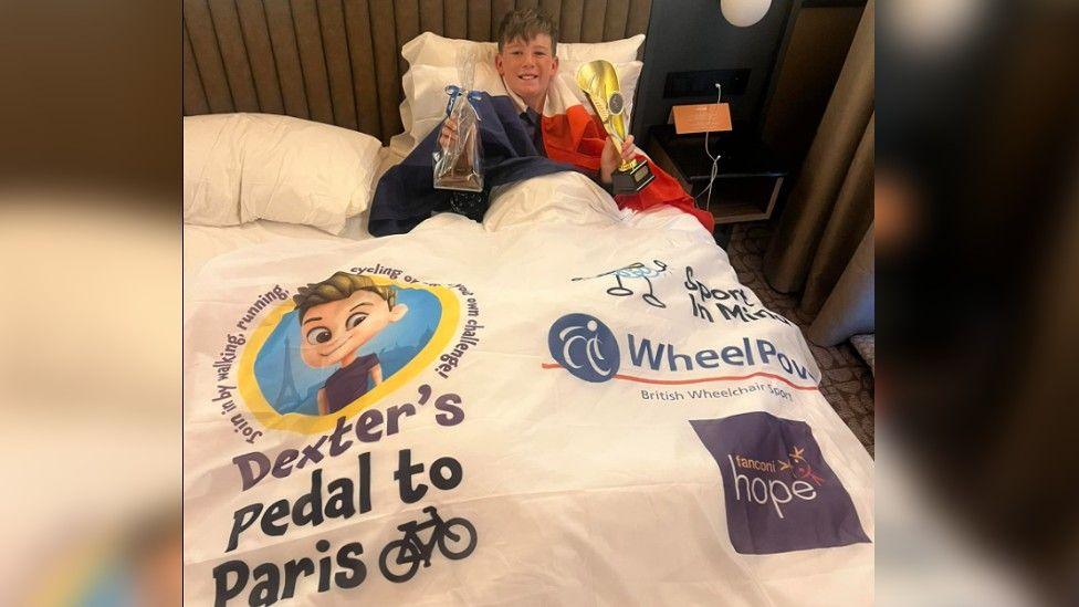 Dexter in bed with a French flag around him, holding a bag of chocolate in his left hand and a gold medal in his right. On the bed is a light weight sheet that says Dexter's Pedal to Paris along with the three charities he raised money for Sport in Mind, WheelPower and Fanconi Hope.