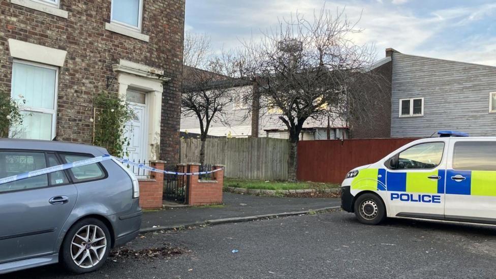 The house in Elswick where the attack took place. The property is an end terrace house made of brown brick. A climbing plant is growing on a trellis next to the white front door. Blue and white police tape has been strung from the property's wall and across a grey car. A police van is parked on the road nearby.