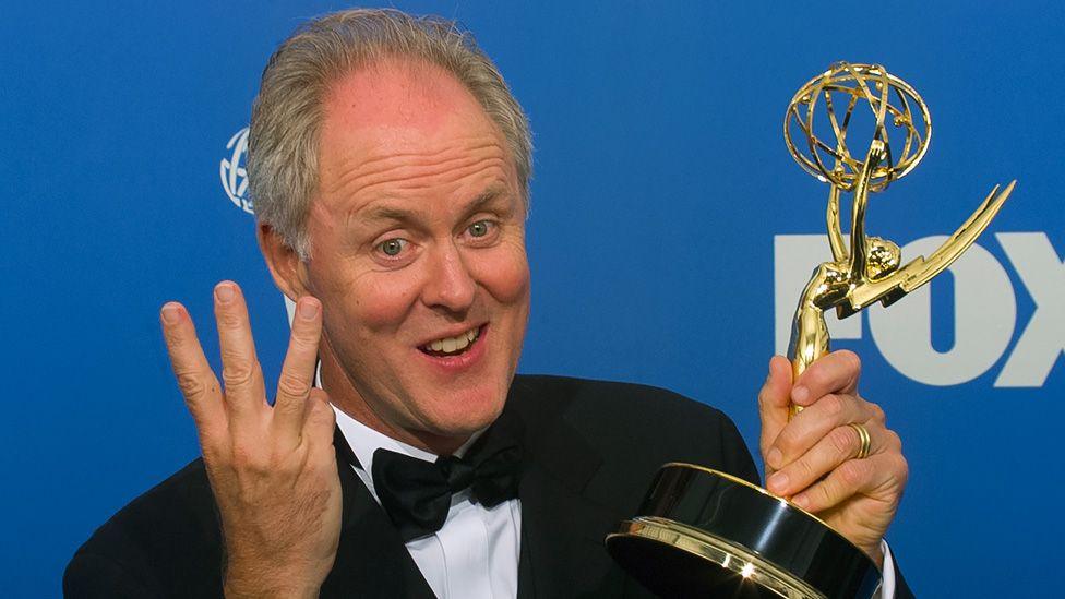 John Lithgow holding up an Emmy trophy with one hand and holding up three fingers on his other hand at the Emmys in 1999