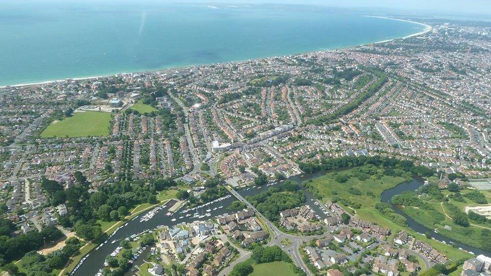 Aerial view over Christchurch, looking towards Bournemouth, Poole and Poole Harbour. The three towns are linked by a long curved sandy beach which runs along the top of the picture with the sea merging with the sky on a misty horizon. Directly below the photographer is the meandering river Avon.
