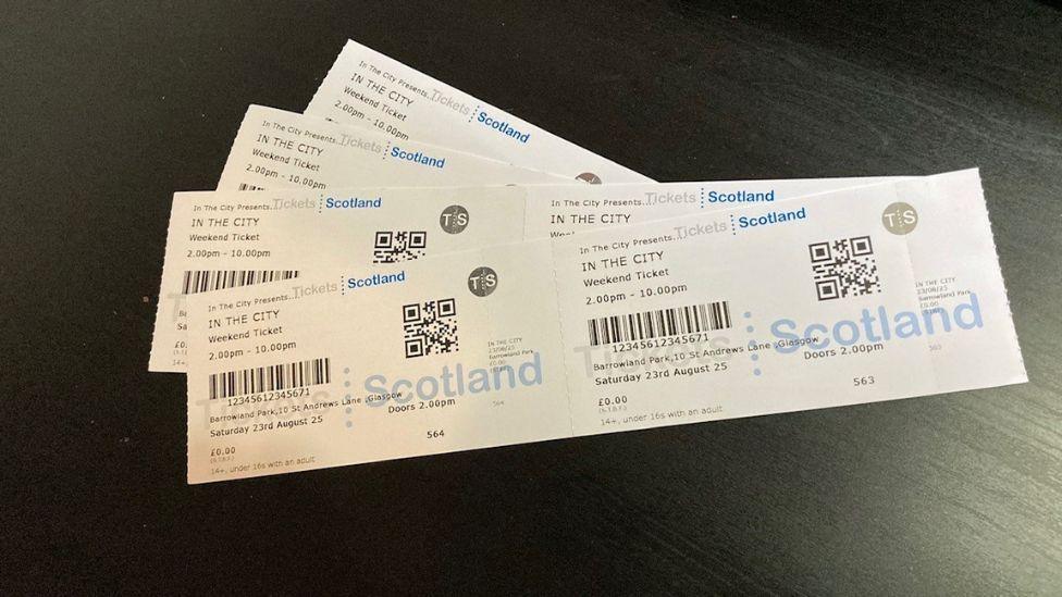 Four white In The City festival tickets on a black wooden surface. The tickets have black text, barcodes and blue logos on them.