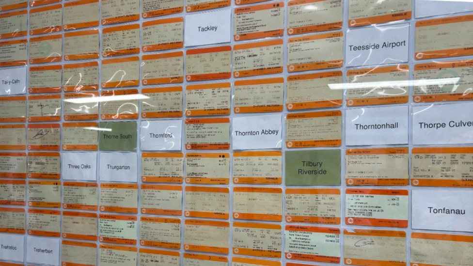 A wall of bright orange rail tickets, with white pieces of paper with the names of railway stations that the collection does not have tickets for yet