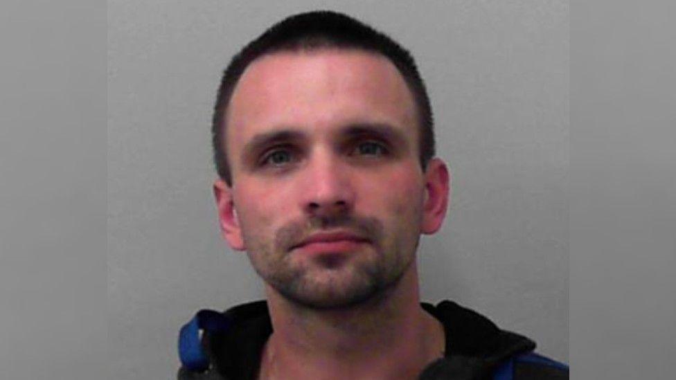 A police mug shot of a slim, white man in his late 30s with close cropped hair and stubble. 