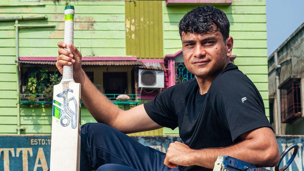 Adnan Miakhel in a publicity shot from Freddie Flintoff's Field of Dreams holding a cricket bat in front of a wooden building painted lime green and wearing navy sports clothes