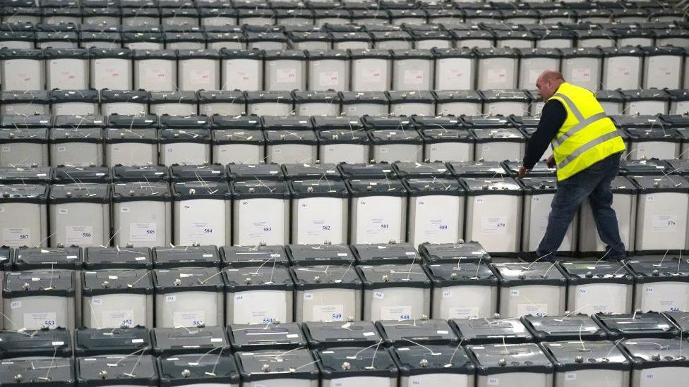 A man in a yellow hi-vis jacket is walking among a sea of grey ballot boxes, checking the lids are secure.