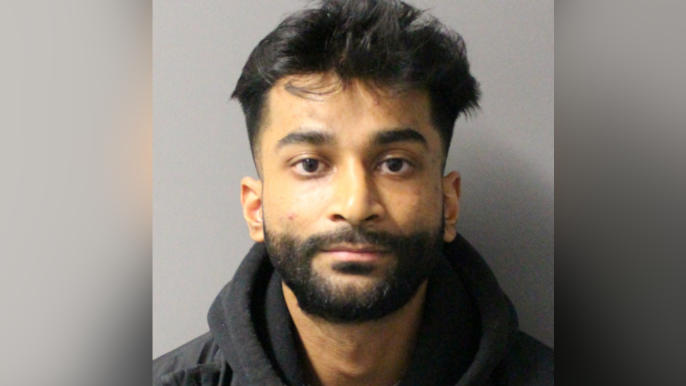A man looks at the camera for a police custody photo. He has a dark brown beard and brown hair and is wearing a black hoodie