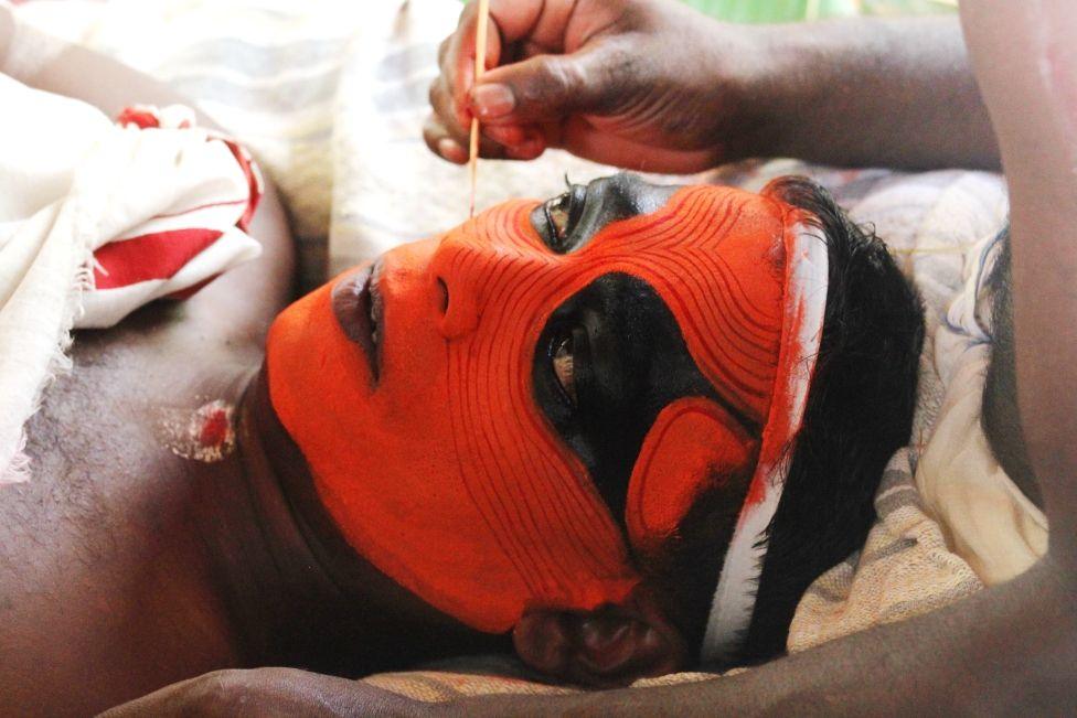 Someone laying on their back and having face paint intricately applied by the hand of another person (unseen)