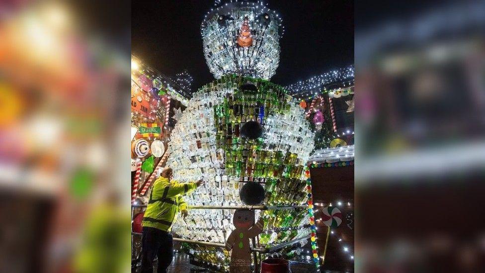 A snowman at night sculpted from 2,500 wine bottles, both clear and green, with Matt Walton dwarfed beside it