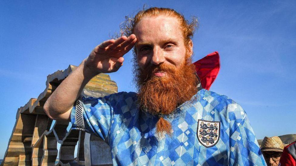 Russ Cook, a red-headed man with a long beard, wearing a football shirt, does a salute to the camera. The sky behind him is bright blue.
