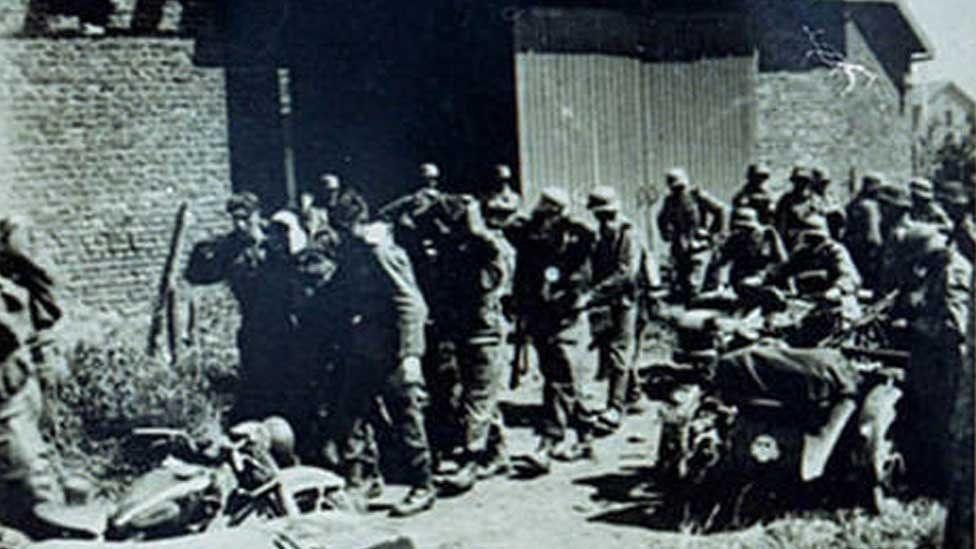 A blurry black and white photo taken in 1940 showing British soldiers surrendering to SS soldiers. There is a barn behind them and two of the British soldiers are holding up a third. A much larger group of SS soldiers are surrounding them, holding guns or sitting on their motorcycles.