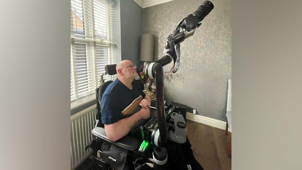 Mr Newcombe sitting in an electric wheelchair behind a long robotic arm with a camera attached to the end