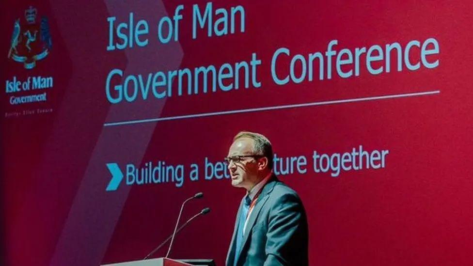 Alfred Cannan wearing glasses while giving a speech with a red presentation in the background with the words government conference, building a better future together written on it..