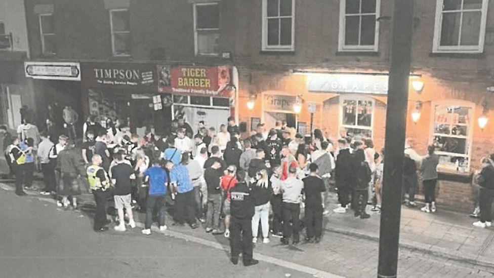 A CCTV image showing a large group of people standing on the pavement outside the pub, surrounded by police officers