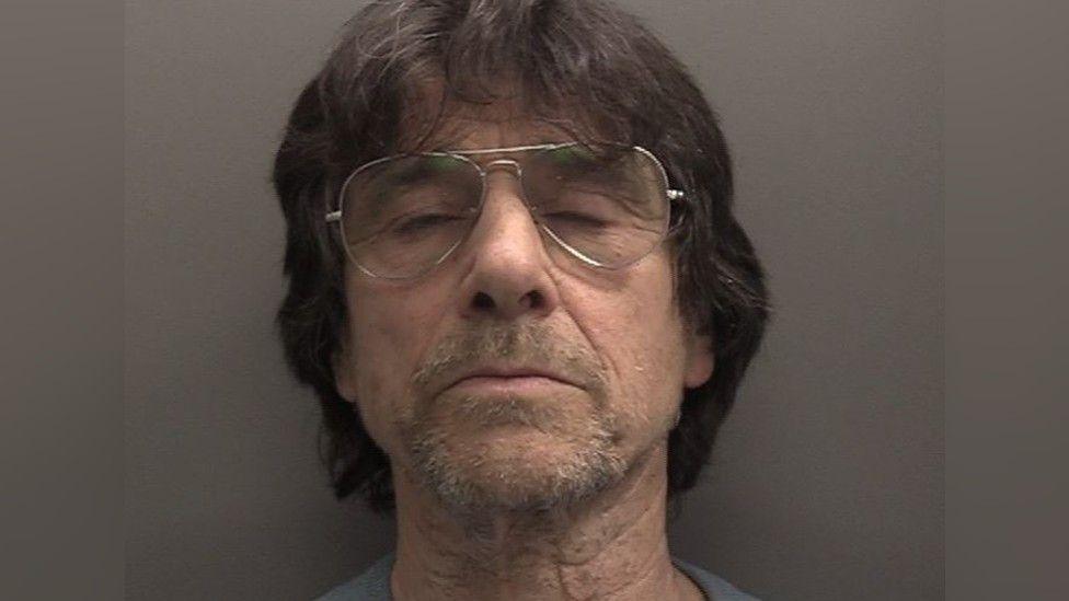 Police mugshot of defendant with eyes closed, shoulder length dark hair, glasses and a goatee 