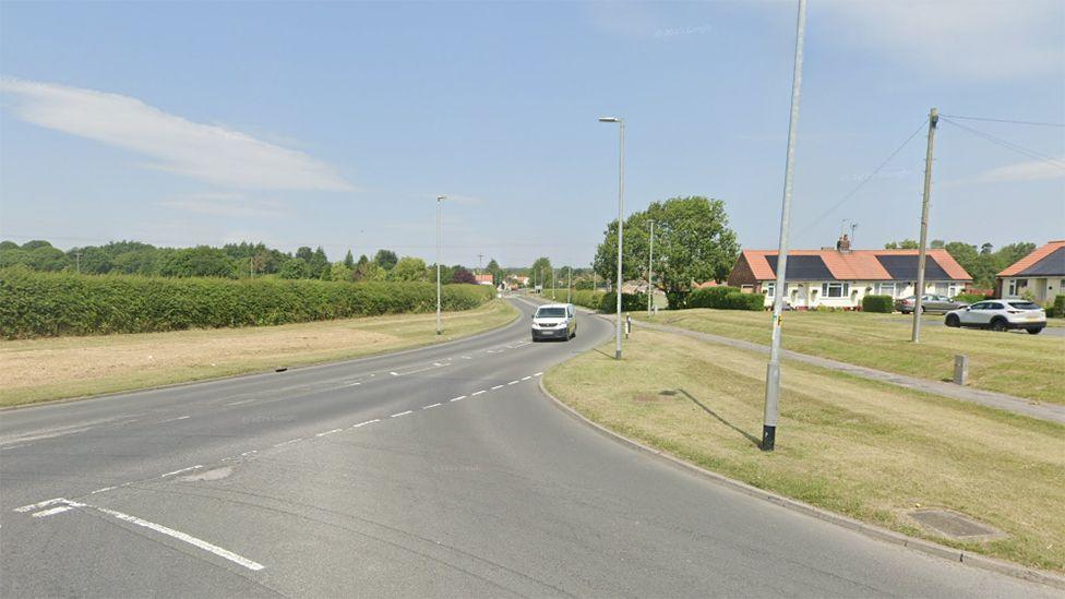 The junction of the A614 and Station Road in Bainton a solitary white van drives towards the junction which is surrounded by grass verges and street lights. In the distance is a hedge and houses
