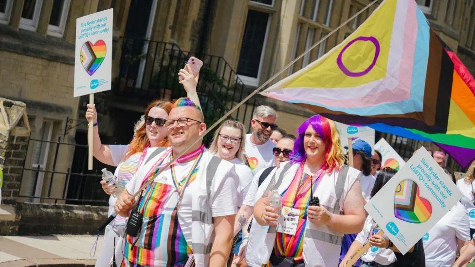 Photo of a Pride event in Peterborough. There is a crowd of people in white T-shirts with rainbow colours in the centre. They are carrying signs reading "Sue Ryder stands with our LGBTQ+ community" as well as a large Progress Pride Flag. Two members of the crowd at the forefront of the image have multicoloured hair. There are buildings in the background. 