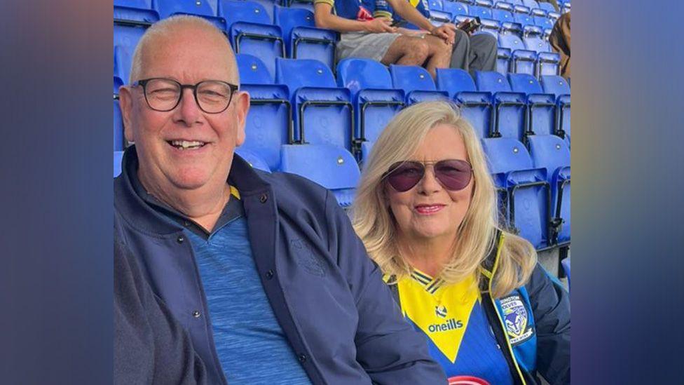 Ged has short grey hair and is wearing dark rimmed glasses and a blue jacket with a blue t-shirt. Jean has long blonde hair and is wearing sunglasses and a Warrington Wolves top and jacket. They are sat in a stand of blue seats with other rugby league fans sat behind them.