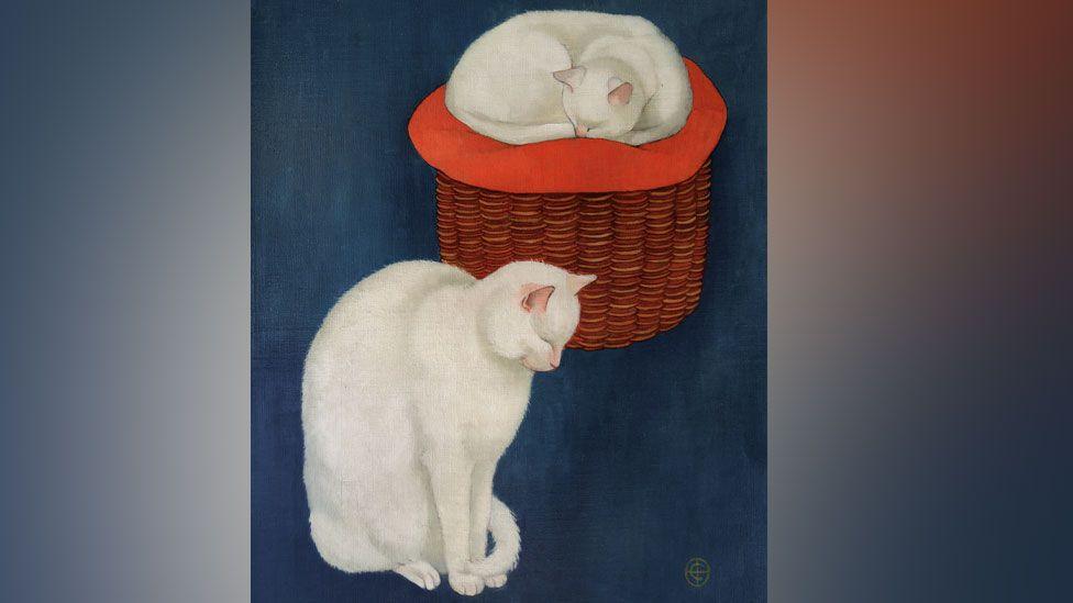 A picture by C Brooke showing two white cats against a deep blue background. The cat on the left is sitting up, with its head down and its tail around its front paws. The second cat is behind it to the right and is curled up on a red cushion on top of a wicker basket. 