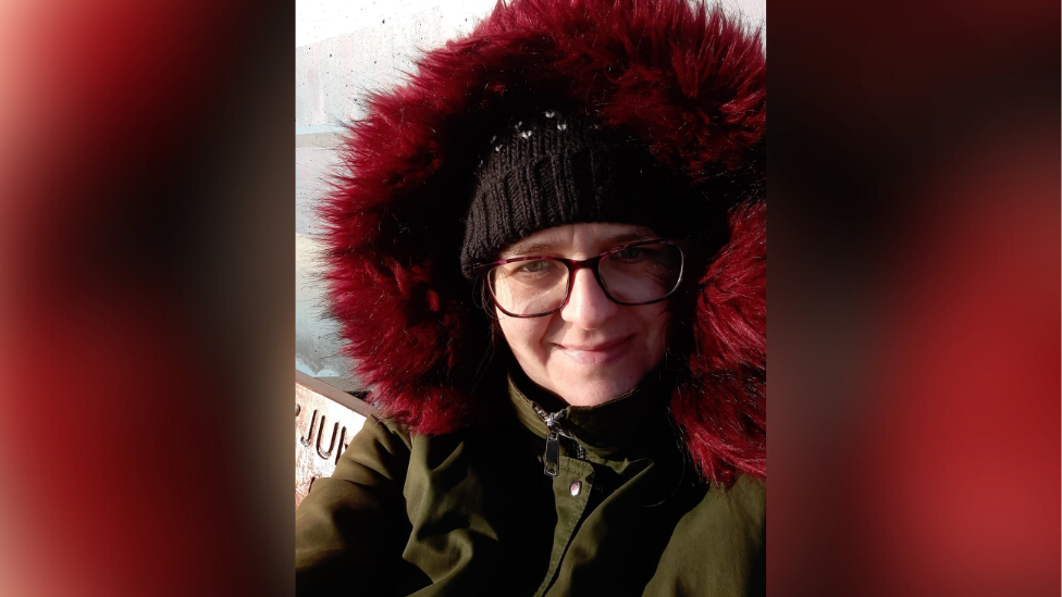 Sarah Young is wearing a green winter coat which has a large, fluffy, red wine coloured hood. She wears a black woollen hat and square framed glasses. She is smiling at the camera and taking a photo of herself in portrait.  