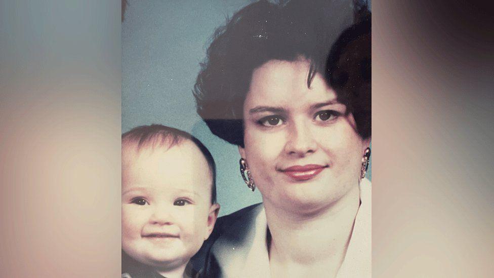 A portrait showing Emily as a baby pictured with her mum Karen, who has black hair, earrings and red lipstick. 