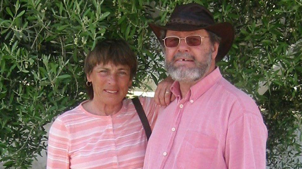 Mary and Noël Hansen, with a bush behind them, posing for a holiday photograph. She has short brown hair; he is wearing a small black stetson hat.