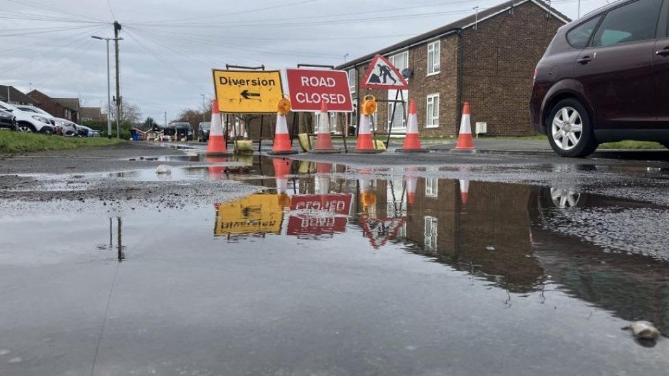 Large puddles of water on the road with signs saying there is a diversion and the road is closed. There are homes and cars parked in the distance