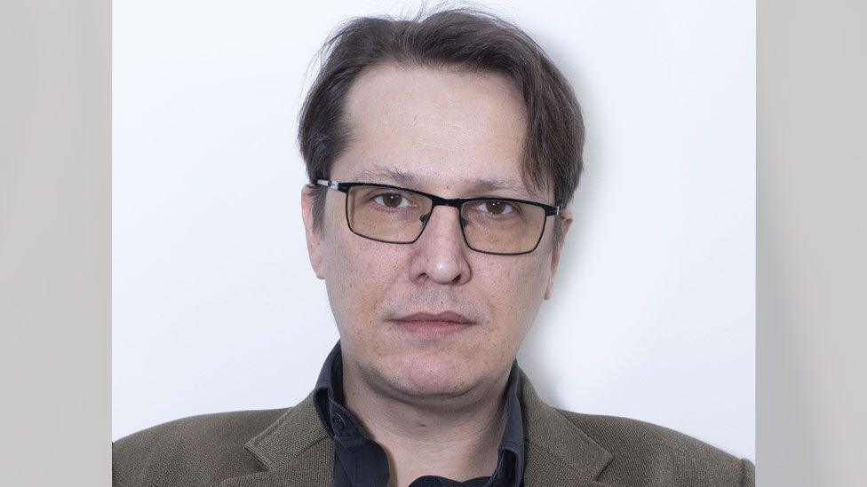 A head and shoulders shot of Stephen O'Grady looking directly down the camera. He is standing against a white background and is wearing a brown jacket over a dark top. He is clean-shaven and is wearing glasses with black, metal, rectangular frames. His dark brown hair is parted on one side.