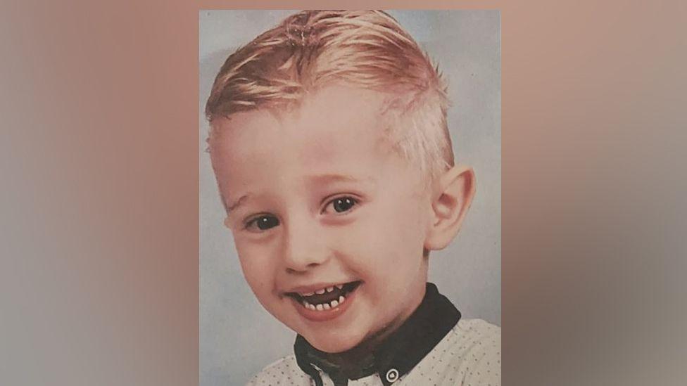 Freddie smiles at the camera as he is professionally photographed. He has blond hair and has a joyful smile at the camera. He is wearing a navy collar shirt.