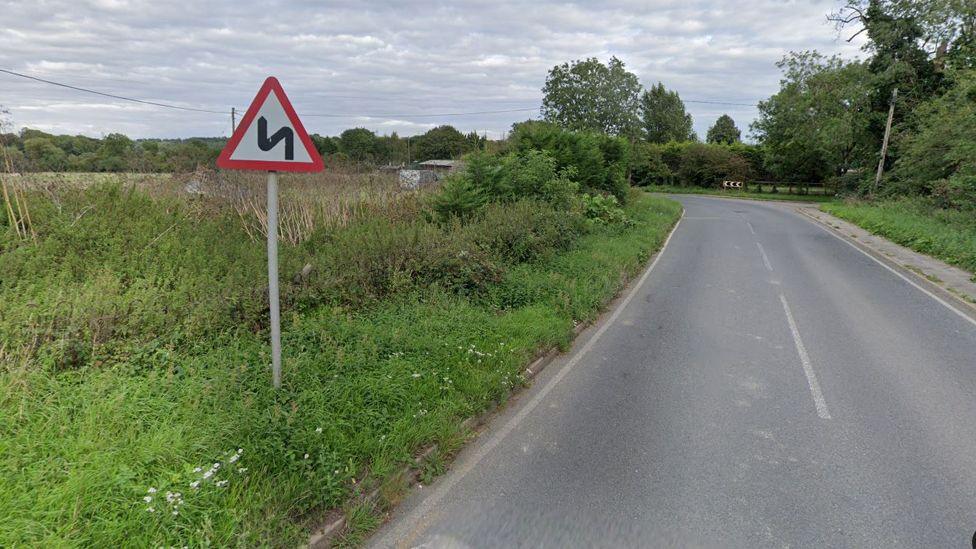 A country lane going into a bend, with a triangle warning sign showing a bendy road. There is grass and shrubbery either side of the road, and on the right side you can see a black-and-white chevron sign next to a small junction.