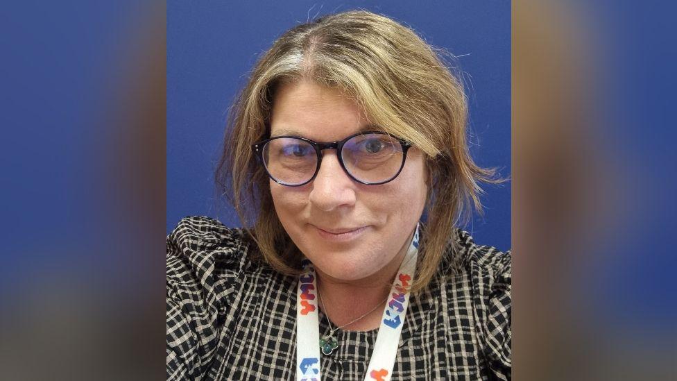A selfie of a woman with neck-length dark blonde hair and glasses. She is wearing a black and white check top and has a lanyard round her neck marked "YMCA".