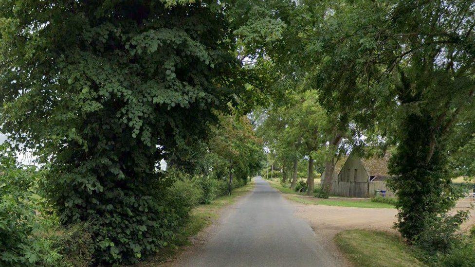 Gote Lane is a narrow, rural road surrounded by trees and shrubs. There is a small, old single-storey building to one side and a dusty track