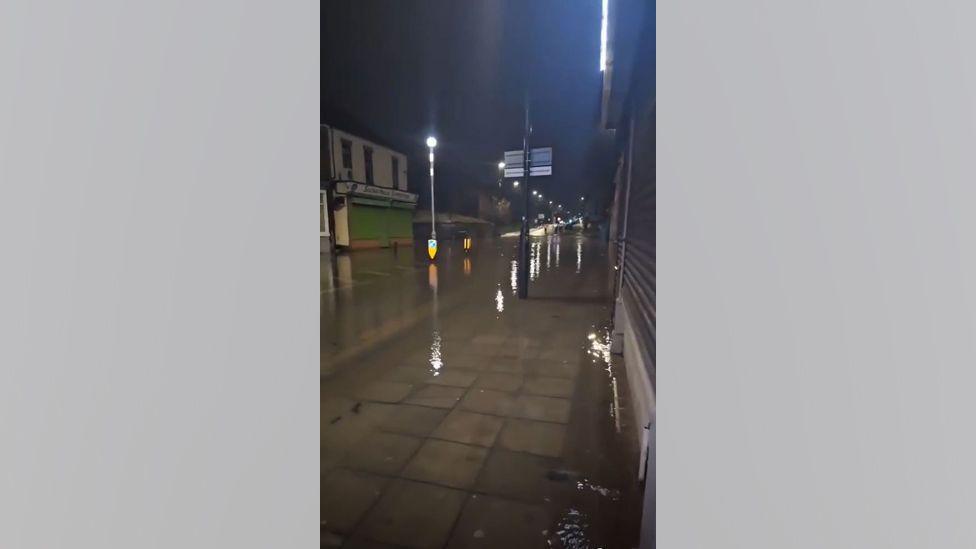 Chester Road following the burst water main. Water has flooded the road and pavement. Light from traffic lights is reflected in the water.