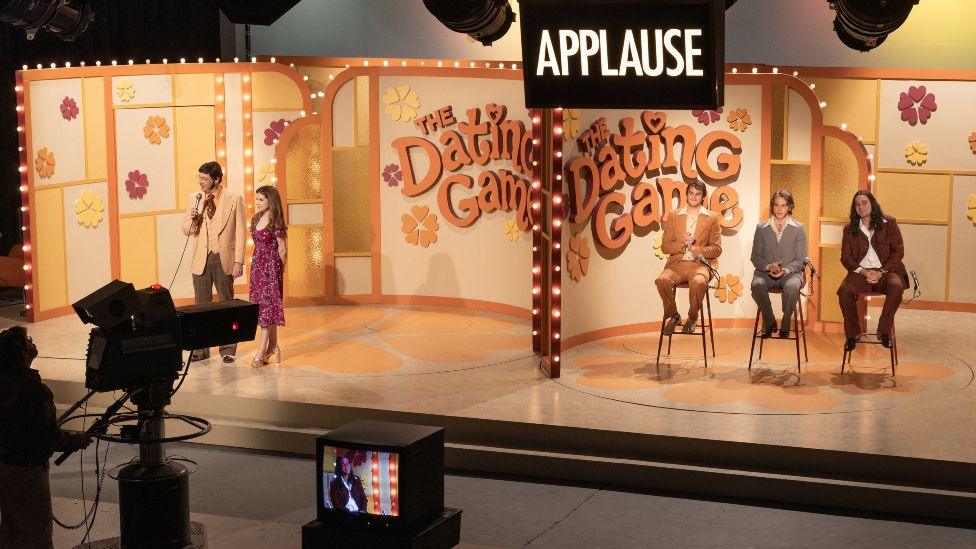 Film still from Woman of the Hour showing contestants on The Dating Game. It shows the host and Anna Kendrick's character on the left hand side. On the right hand side of the TV set are three male contestants sitting on stools. The two sides of the stage are separated by a screen. In the background is a sign sating The Dating Game.