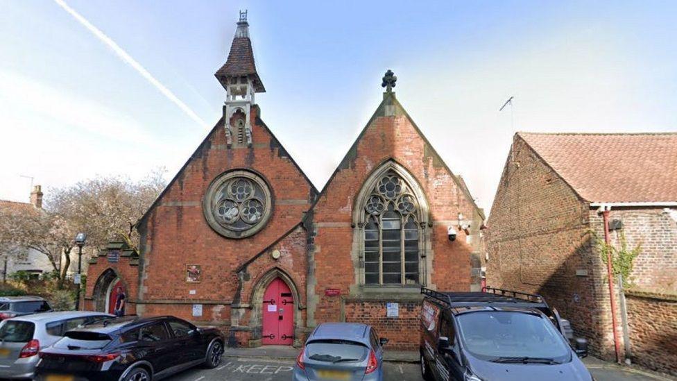 Beverley Minster Parish Hall before the bell tower was removed. There are cars parked in front of the red brick building.