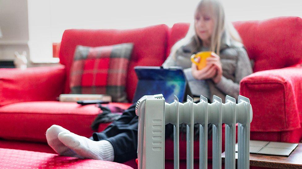 Stock photo shows an elderly woman sits on the sofa with a cup of tea next to a heater, trying to keep warm