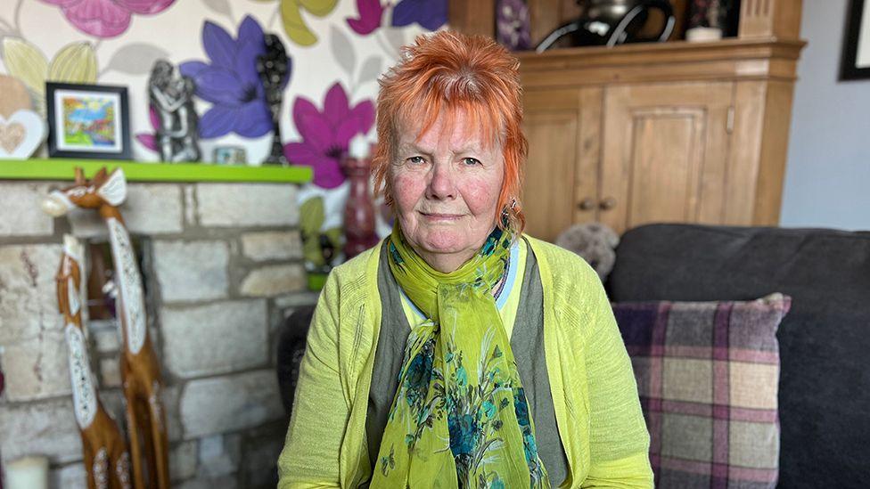 Jenny Thurlow has orange hair and wears a green top and scarf as she sits in her home in front of a stone fireplace