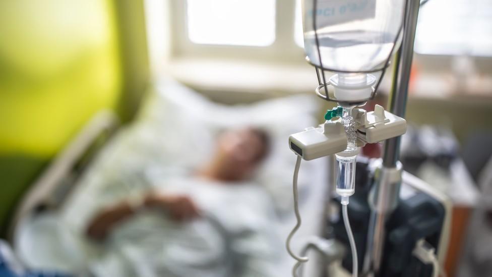 A stock image showing an out-of-focus patient lying in a hospital bed. In the foreground and in focus is a drip.