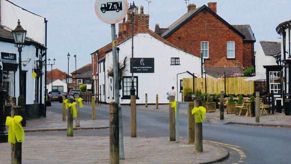 Yellow ribbons tied to wooden bollards in a village street with black and white buildings in the background.