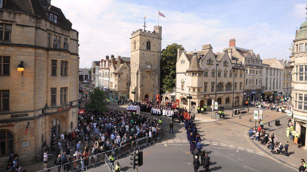 With the sun shining on Oxford's medieval Carfax Tower, several dozen people have gathered behind temporary metal barriers to watch a ceremony.  A procession of people in suits can be seen and some choristers wearing traditional white dress 