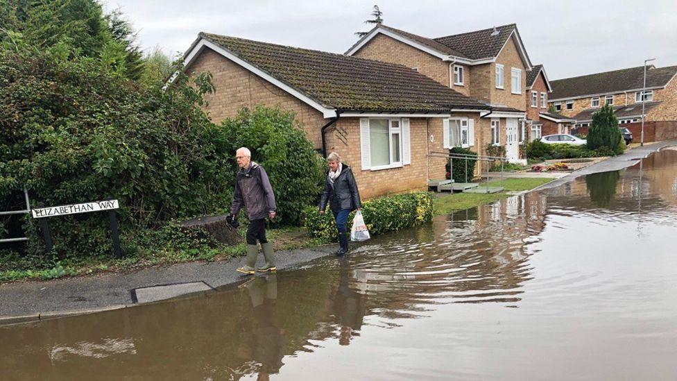 A man and a woman walking past floodwater on a street, with water overflowing the pavement and running up close to homes. They are wearing wellington boots.